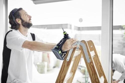En man står på en stege inomhus, håller i en sladdlös borrmaskin och tittar uppåt nära ett fönster, möjligen i färd med att förbereda en reparation eller installation.