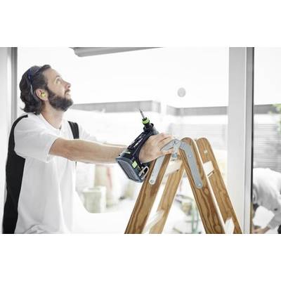 En man står på en stege inomhus, håller i en sladdlös borrmaskin och tittar uppåt nära ett fönster, möjligen i färd med att förbereda en reparation eller installation.