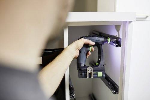 En person som använder en sladdlös skruvdragare för att installera lådskenor inuti ett vitt skåp, med fokus på precision och justering.