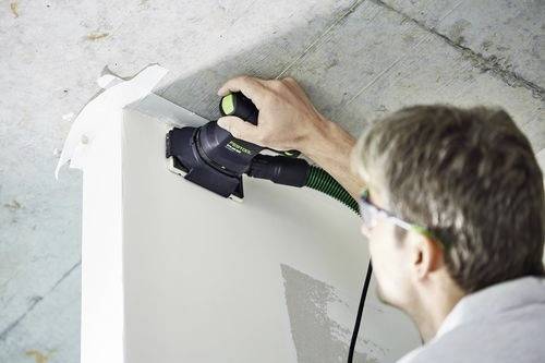 En person använder en elektrisk slipmaskin på ett vitt hörn av en vägg och jämnar ut ytan i ett nyligen konstruerat eller renoverat interiört utrymme.