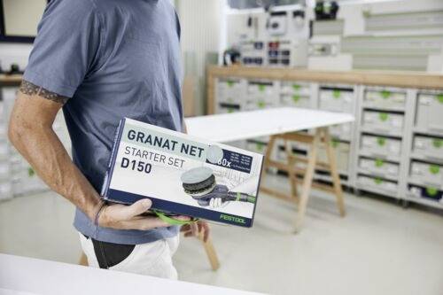 En person håller i en 'Granat Net Starter Set D150' låda i en verkstad. Bakgrunden visar hyllor med förvaringsenheter och ett arbetsbord.
