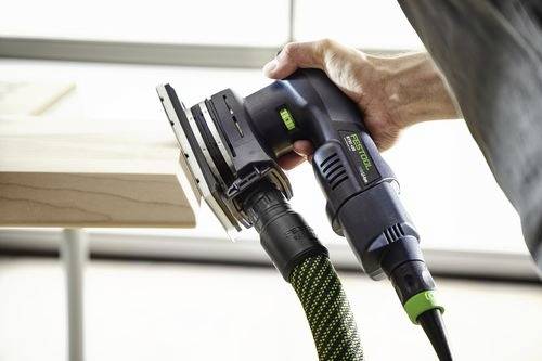 En person använder en elektrisk slipmaskin på en träyta och håller verktyget med en hand. Slipmaskinen är ansluten till en grön och svart slang.