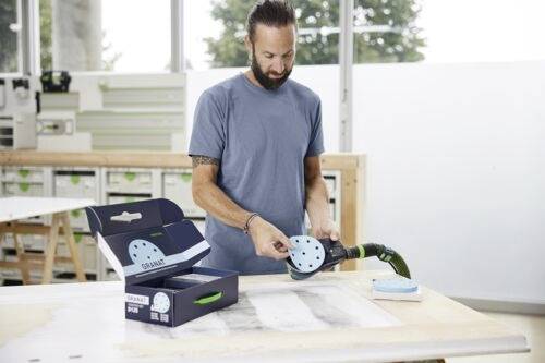 En man i en verkstad som använder ett elektriskt slipverktyg på en arbetsbank, med en låda med slipskivor intill.