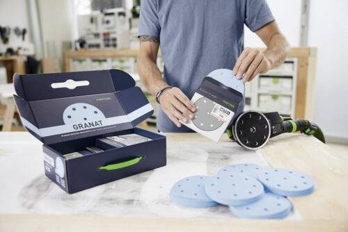 En person i en verkstad håller en cirkulär slipskiva bredvid en maskin. En låda märkt 'Granat' och fler skivor finns på ett bord.