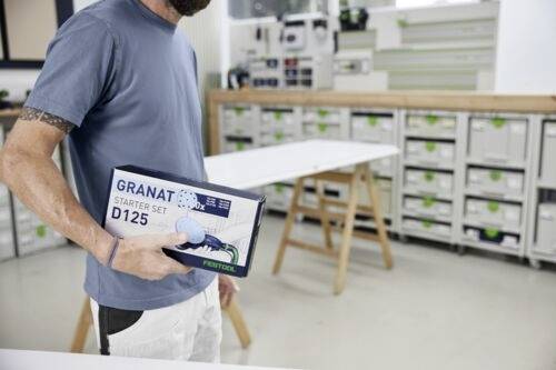 En person i en verkstad håller i en 'GRANAT Starter Set D125' låda. Hyllor med organiserade behållare syns i bakgrunden.