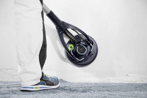 En person använder ett Festool-verktyg med ett roterande huvud för att slipa en vit vägg, iklädd ljusa byxor och sneakers, stående på ett heltäckningsmatta.