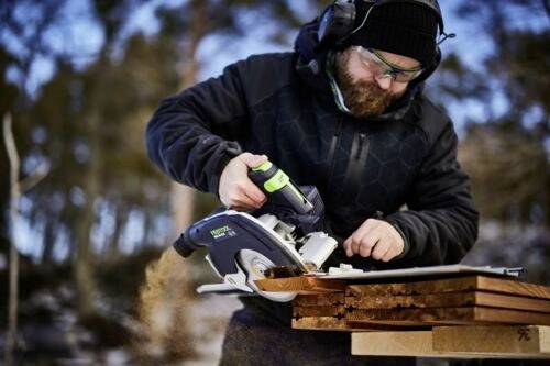 En person som använder en cirkelsåg för att såga träplankor utomhus, iklädd skyddsutrustning med en bakgrund av träd.