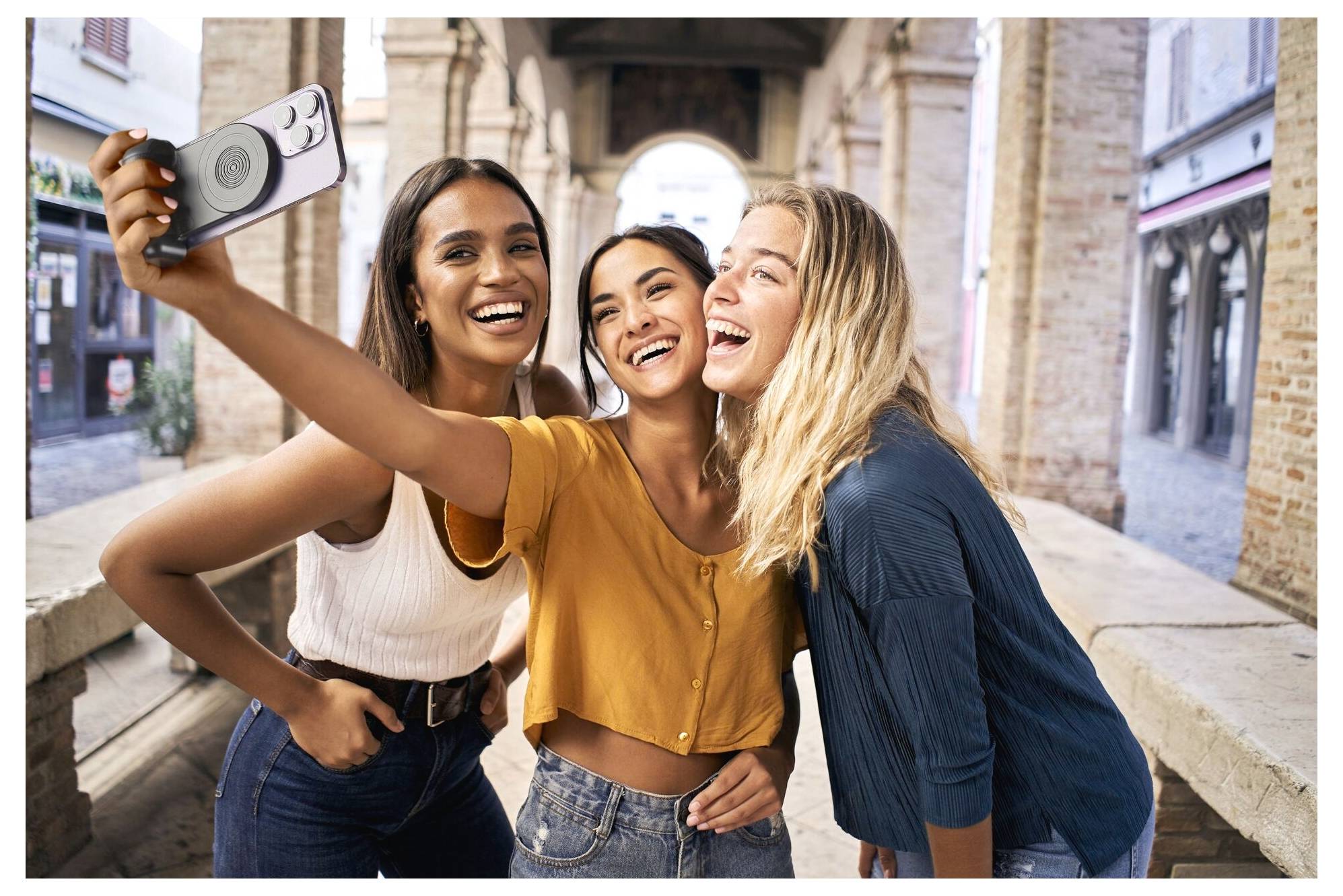 Tre vänner som ler medan de tar en selfie vid en historisk stenbåge i en europeisk gränd.