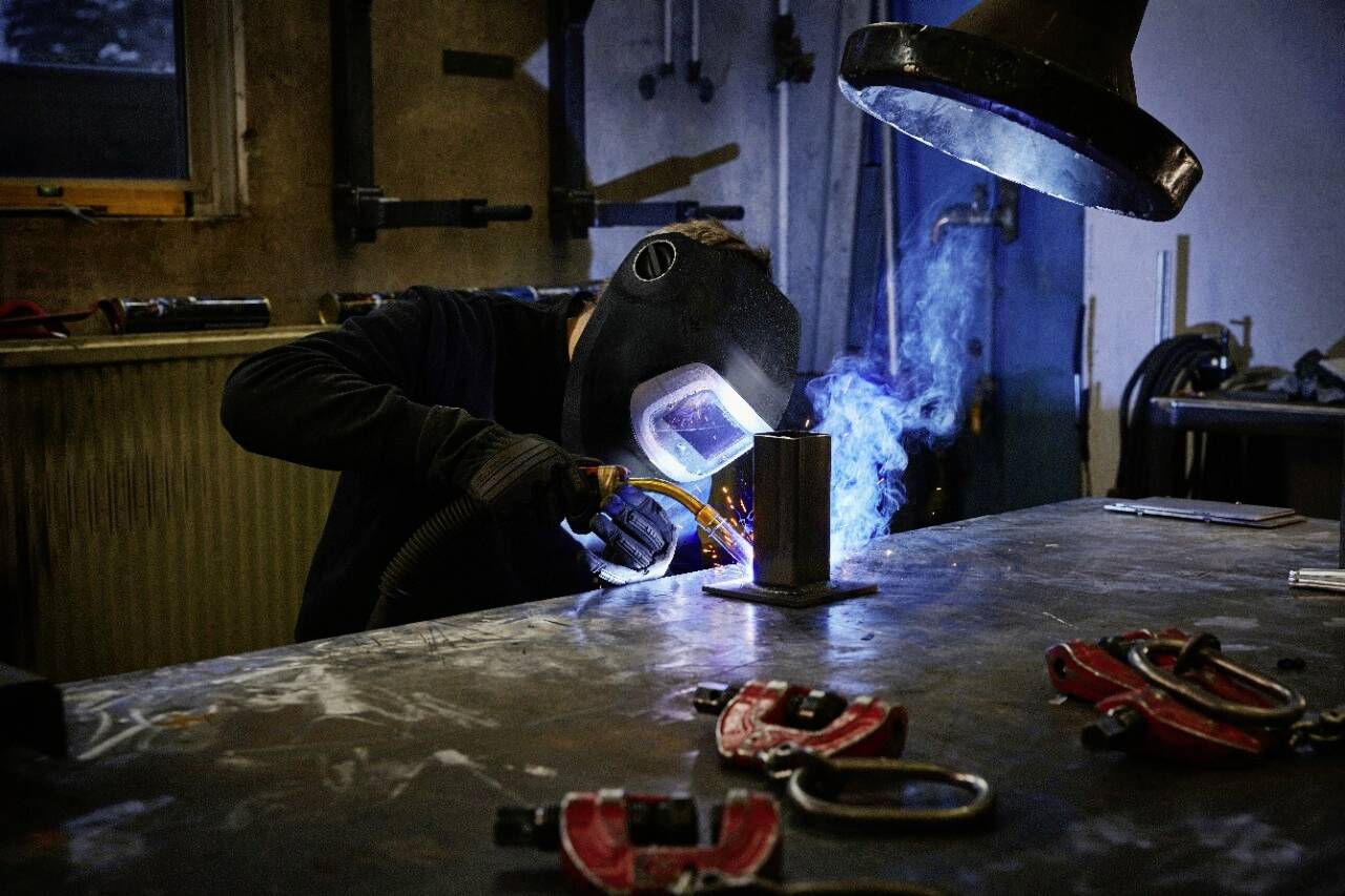 En person med en svetshjälm svetsar metall i en verkstad, omgiven av verktyg och maskiner, med synliga gnistor och rök.