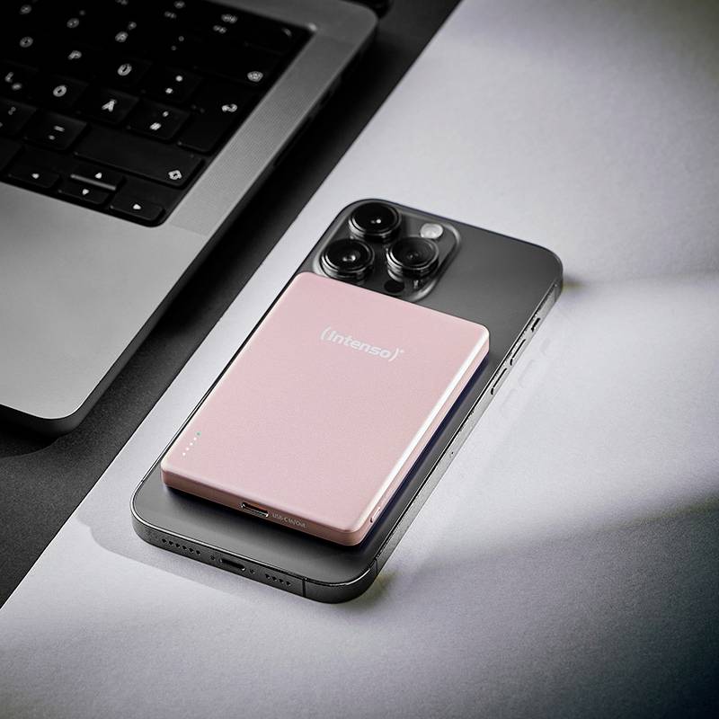 En smartphone, en rosa Intenso-varumärkets powerbank och en delvis synlig laptop på ett skrivbord, vilket antyder en teknisk eller arbetsrelaterad uppställning.