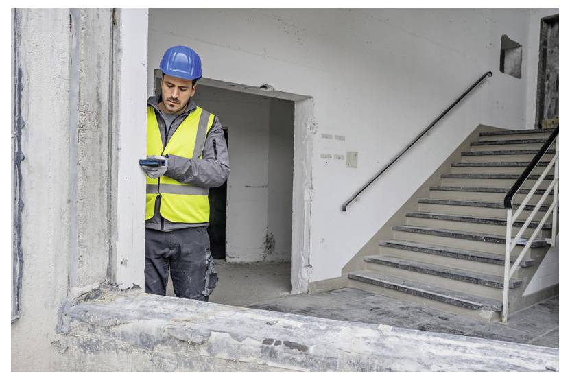 En byggnadsarbetare med säkerhetsväst och hjälm undersöker en enhet i en delvis renoverad byggnad intill en trappa.