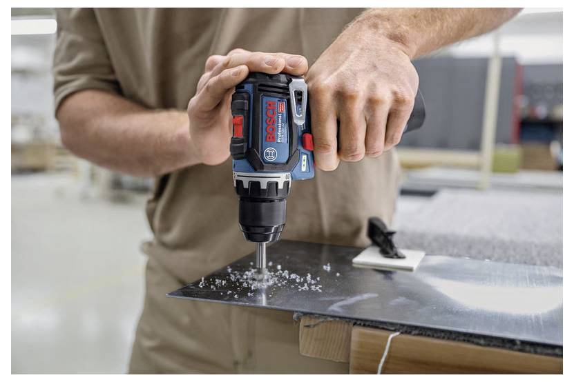 En person använder en Bosch-borr för att skapa ett hål i ett metallark i en verkstad, med metallspån synliga, vilket betonar verktygets precision och kraft.