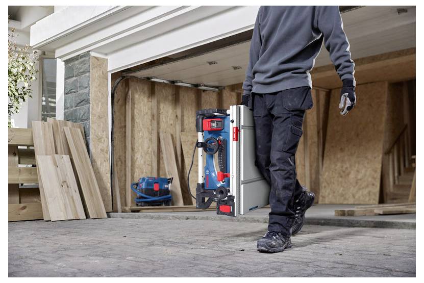 En person i arbetsklädsel bär en blå och röd Bosch-verktygslåda längs en uppfart framför en öppen garage som innehåller träplankor.
