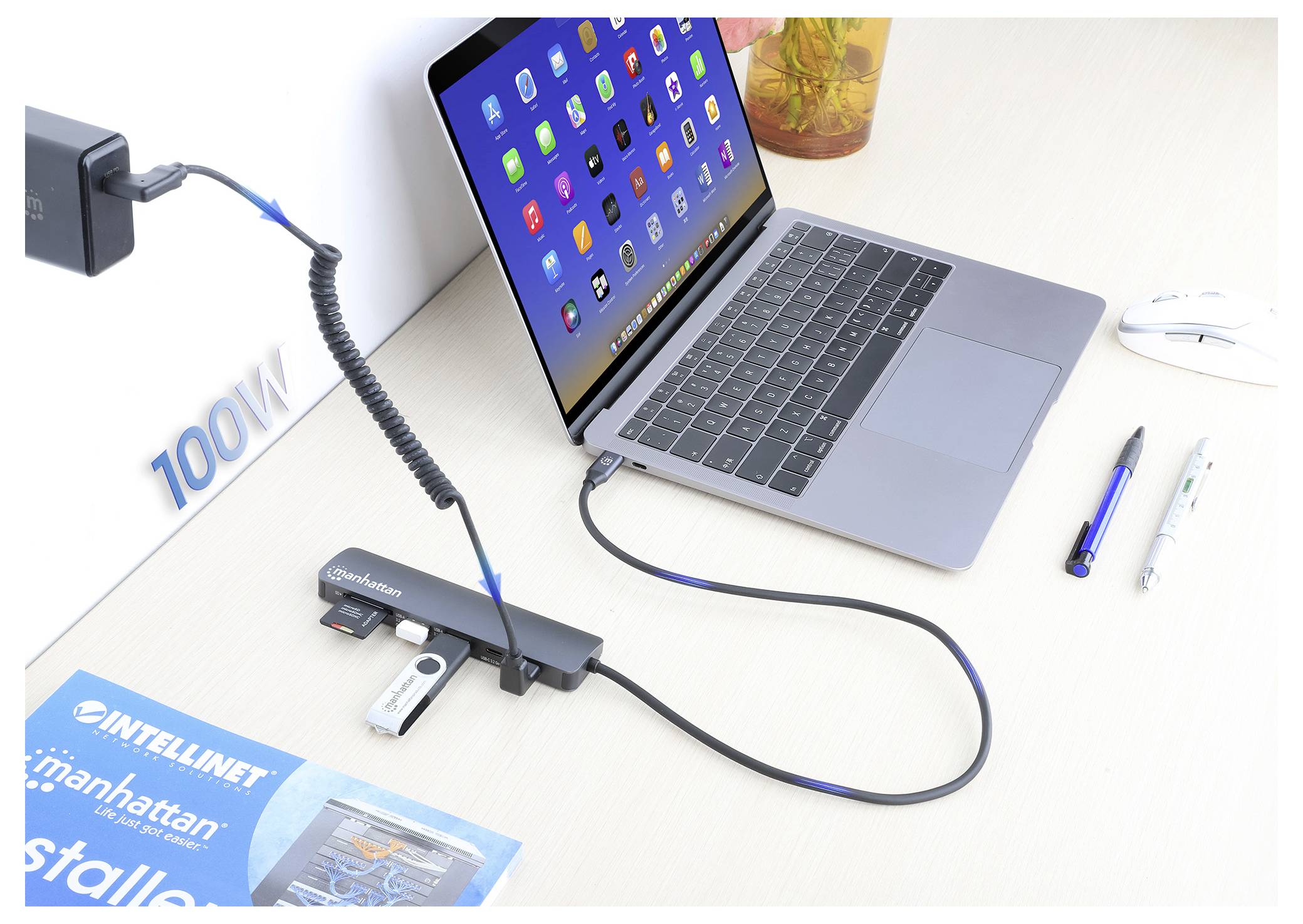 En bärbar dator ansluten till en dockningsstation med flera portar och enheter, inklusive ett USB-minne, på ett skrivbord. Text '100W' synlig.
