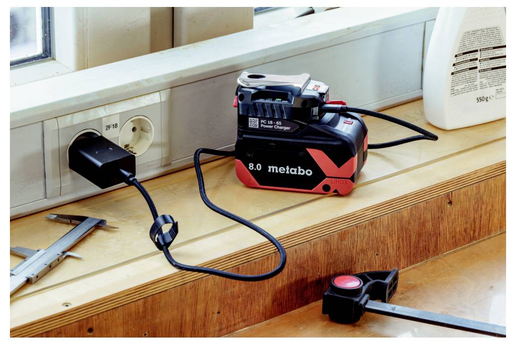 En Metabo batteriladdare inkopplad i ett vägguttag på en fönsterbräda, med ett måttband och ett skjutmått liggande i närheten på en arbetsbank.