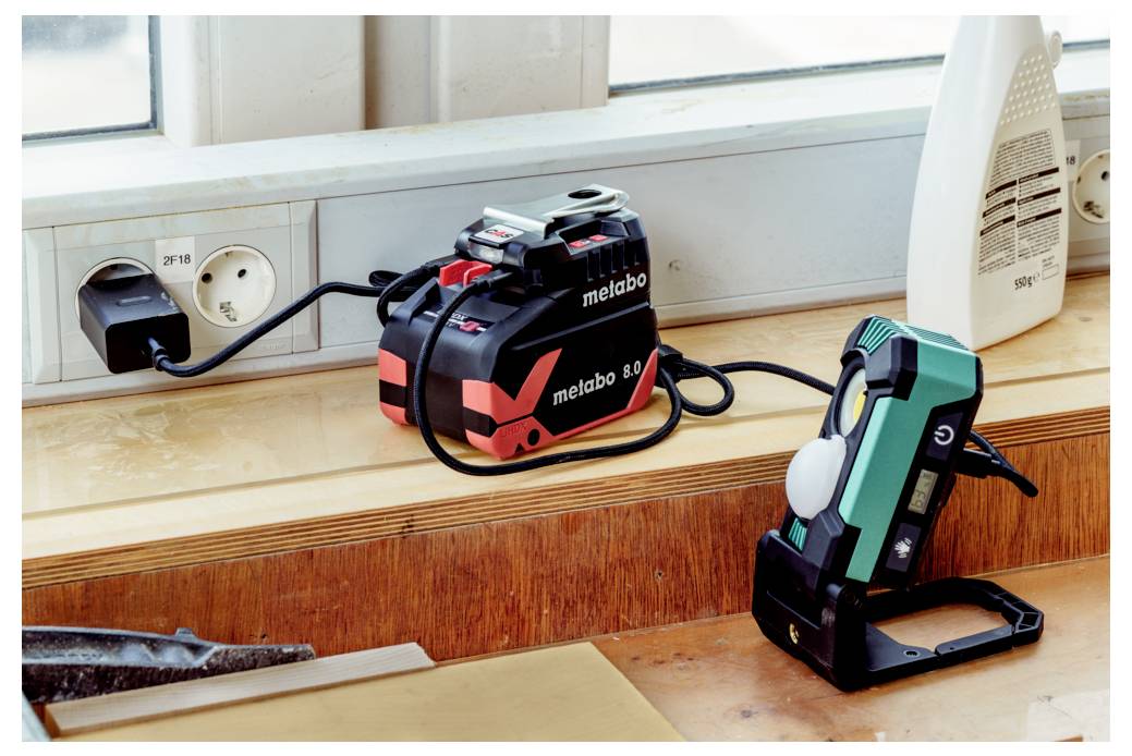 En Metabo batteriladdare inkopplad i ett vägguttag på en fönsterbräda, som laddar ett batteri. En portabel arbetslampa står i närheten.