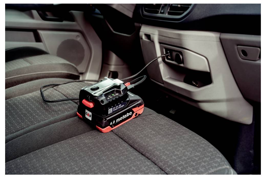 En bilinteriör som visar en Metabo batteriladdare inkopplad i ett eluttag, vilande på ett säte, med synlig armstöd och dörrpanel.