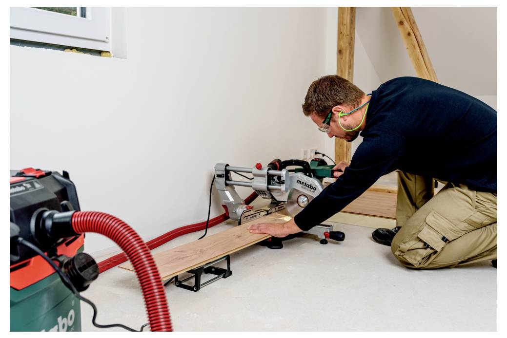 En man med hörselskydd använder en kapsåg för att såga träplankor i en arbetsyta. En dammsugare är ansluten till sågen för dammuppsamling.