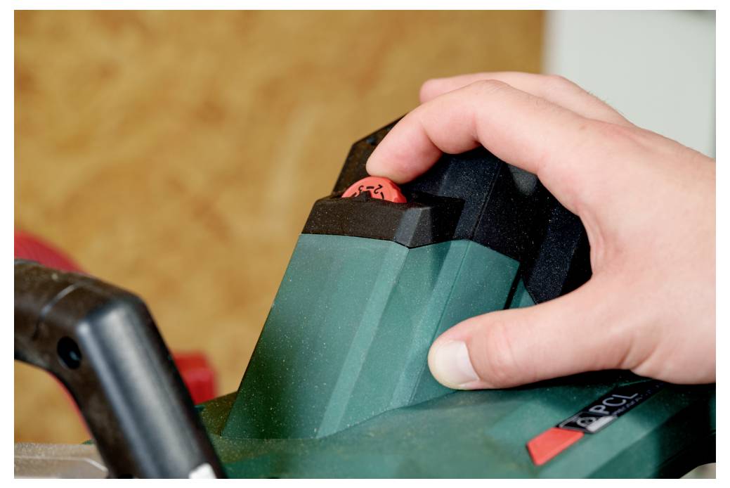 En hand trycker på den röda säkerhetsbrytaren på ett grönt elverktyg, vilket antyder aktivering eller drift i en verkstadsmiljö.