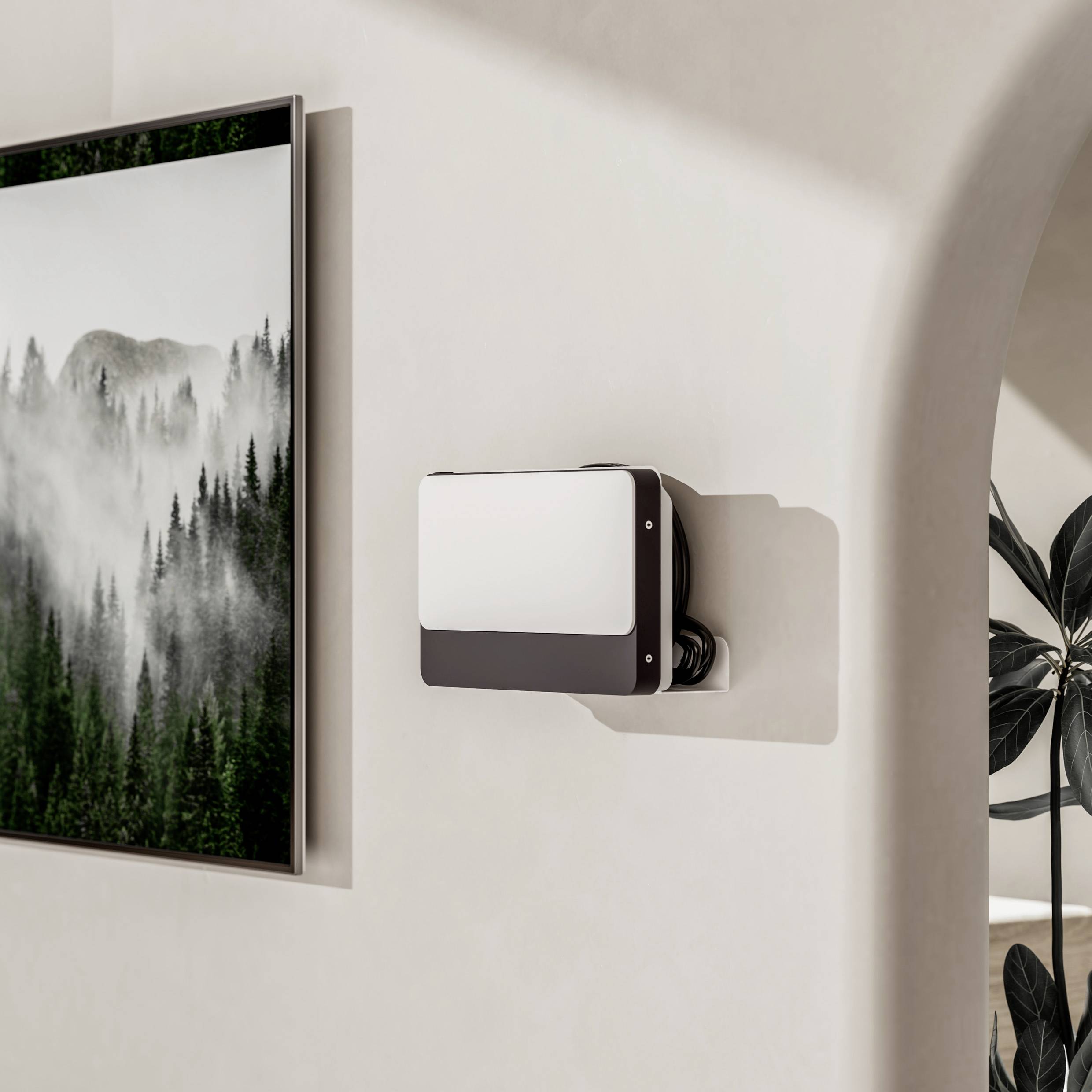 En vit, rektangulär elektronisk enhet är monterad på en krämfärgad vägg bredvid en inramad bild av ett dimmigt skogslandskap.