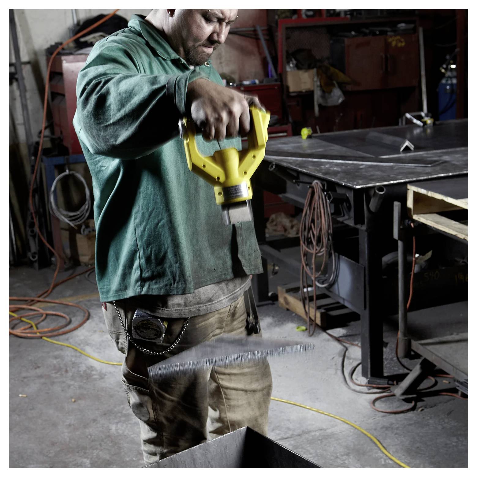 Arbetare i grön tröja klipper metallark med ett elverktyg i en verkstad, omgiven av verktyg och utrustning.