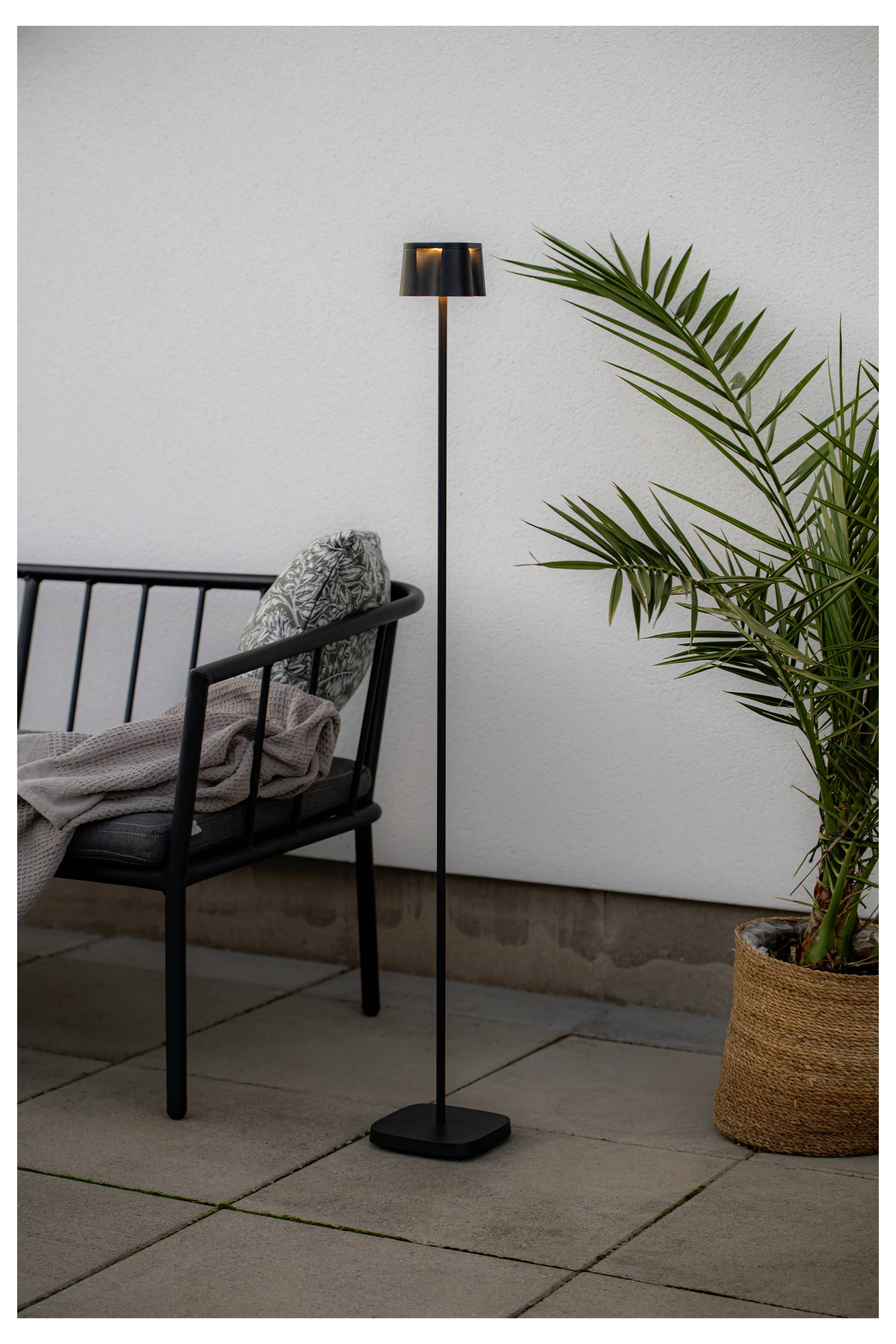En elegant svart golvlampa står bredvid en svart utomhusstol med en kudde och ett påkastat täcke. En krukväxt med palm placeras till höger.