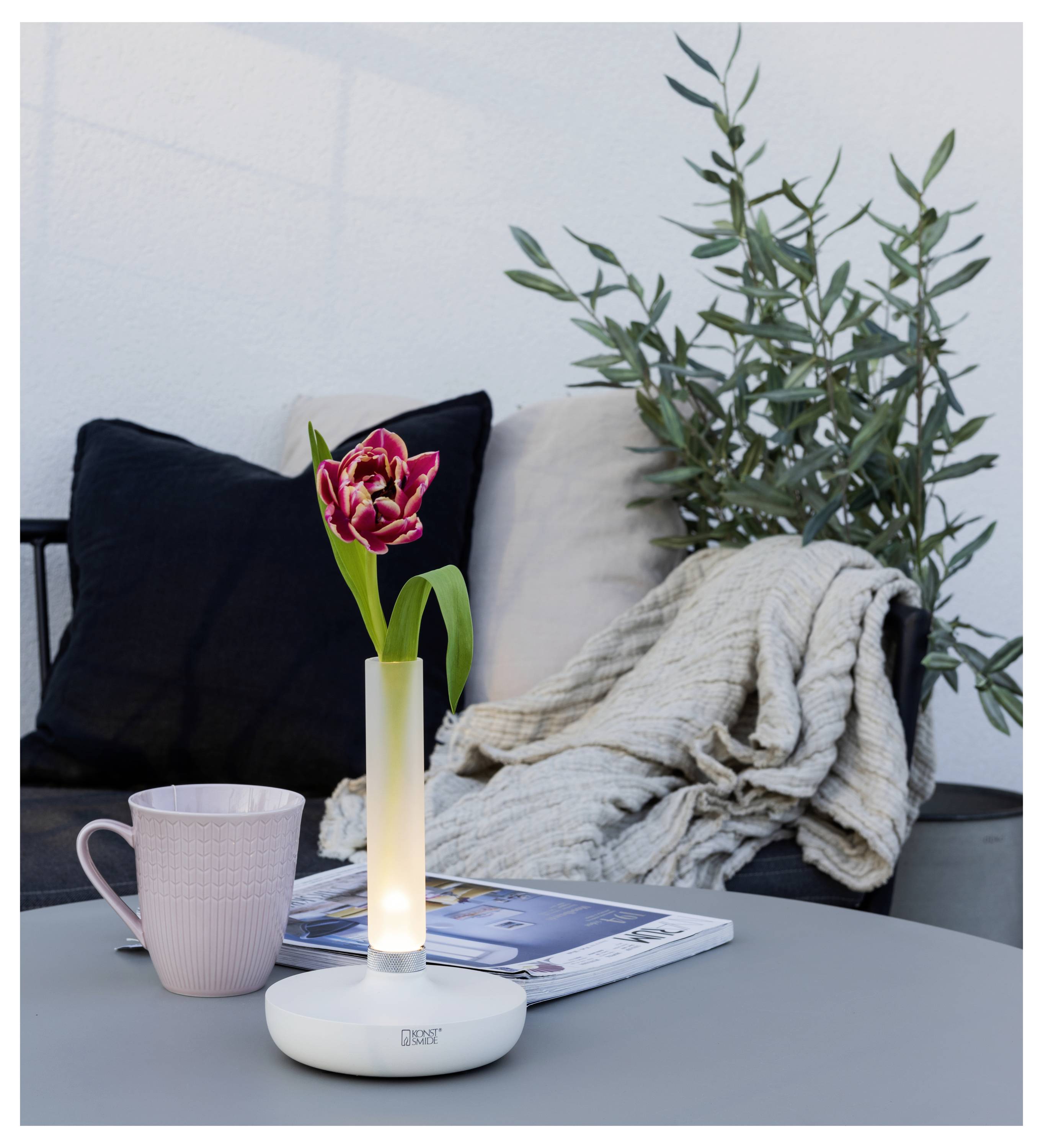 En mysig utomhusmiljö med en glödande lampvas som håller en röd blomma, bredvid en rosa mugg, en tidskrift och en draperad filt på en stol.