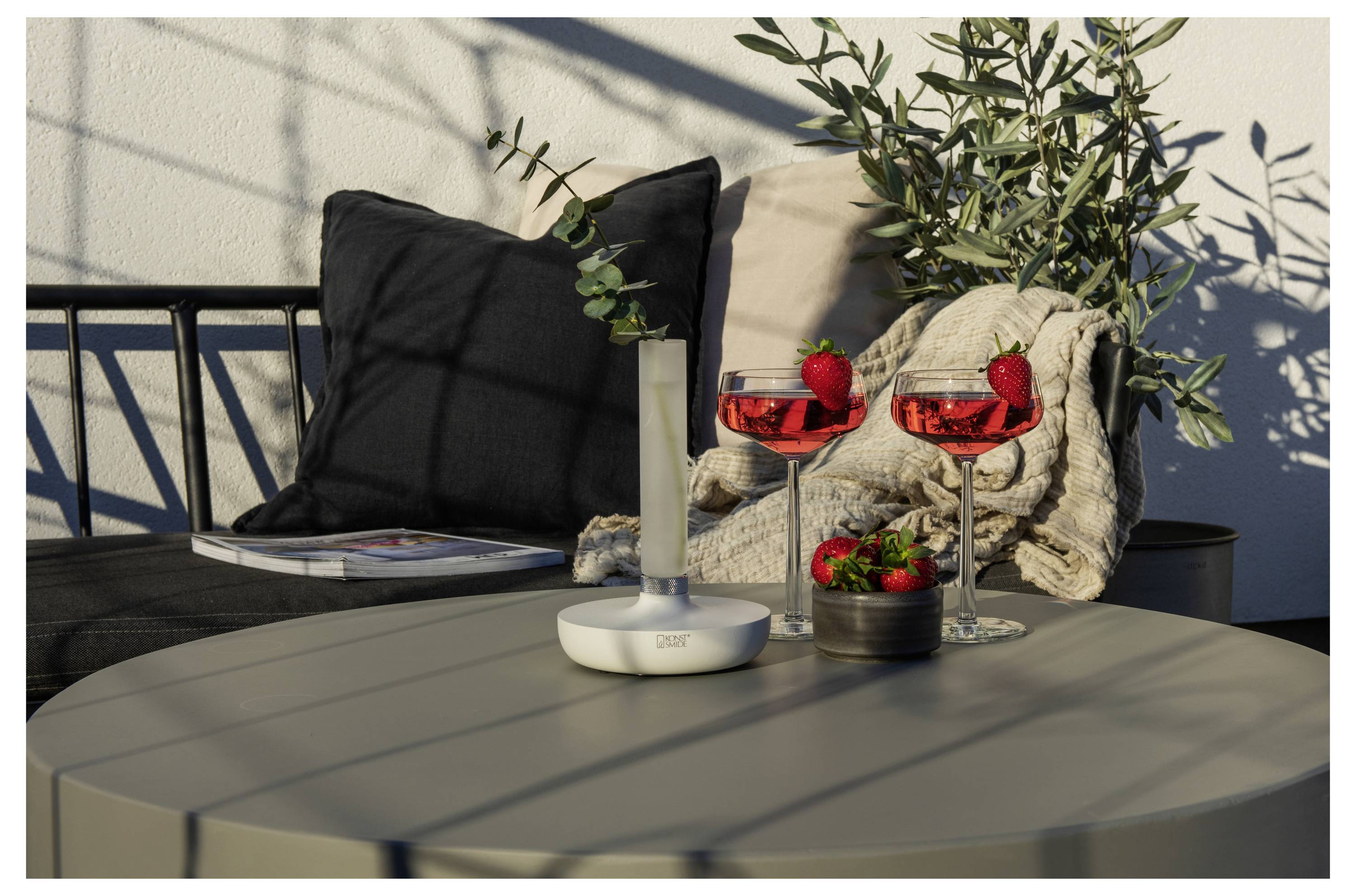 Utomhusmatplats med ett runt bord som har två röda cocktails garnerade med jordgubbar, en vas med en växt och en tidskrift.