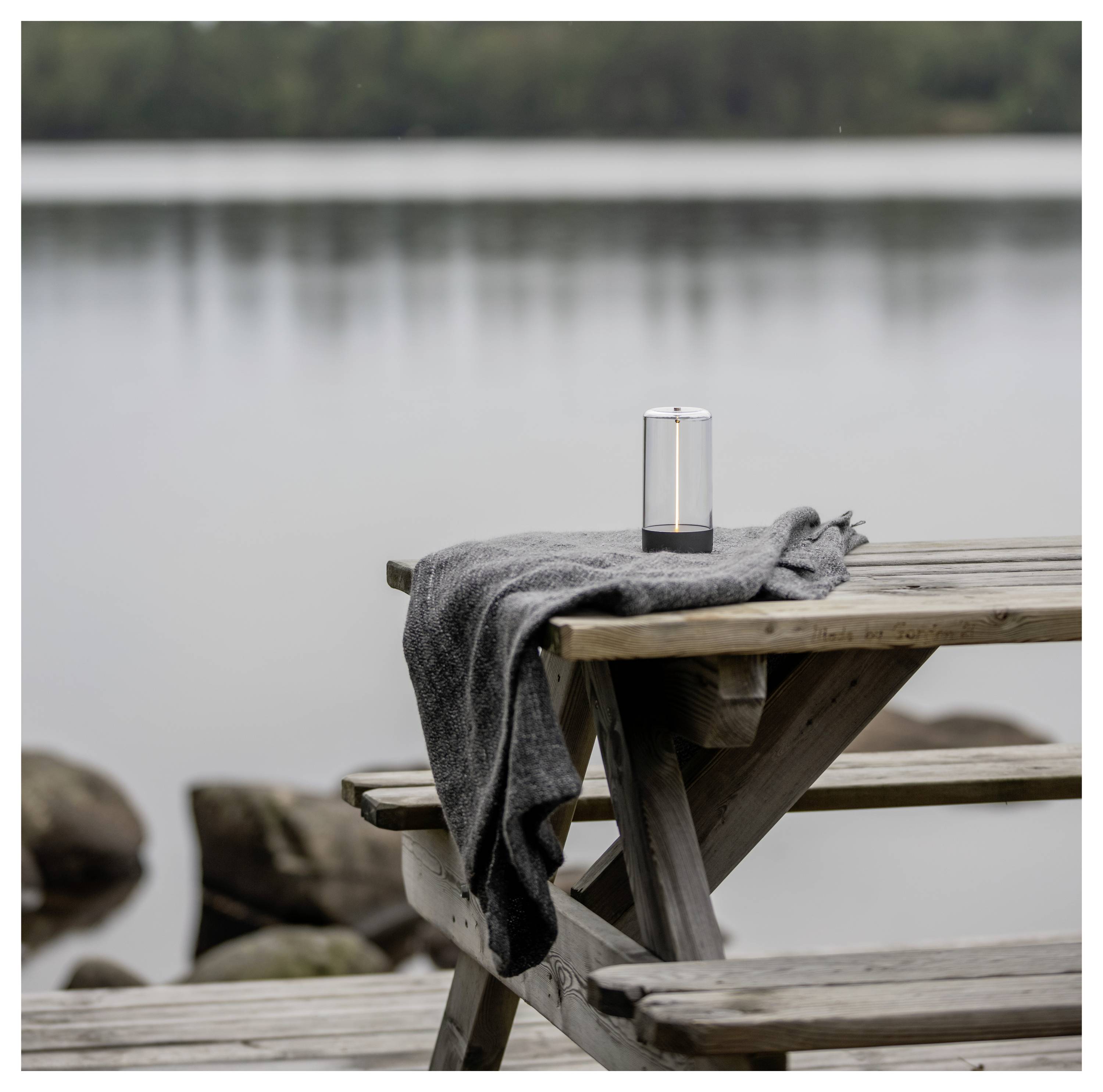Ett trädgångsbord vid en lugn sjö med en mysig grå filt utbredd över bänken och en liten högtalare placerad på bordet.