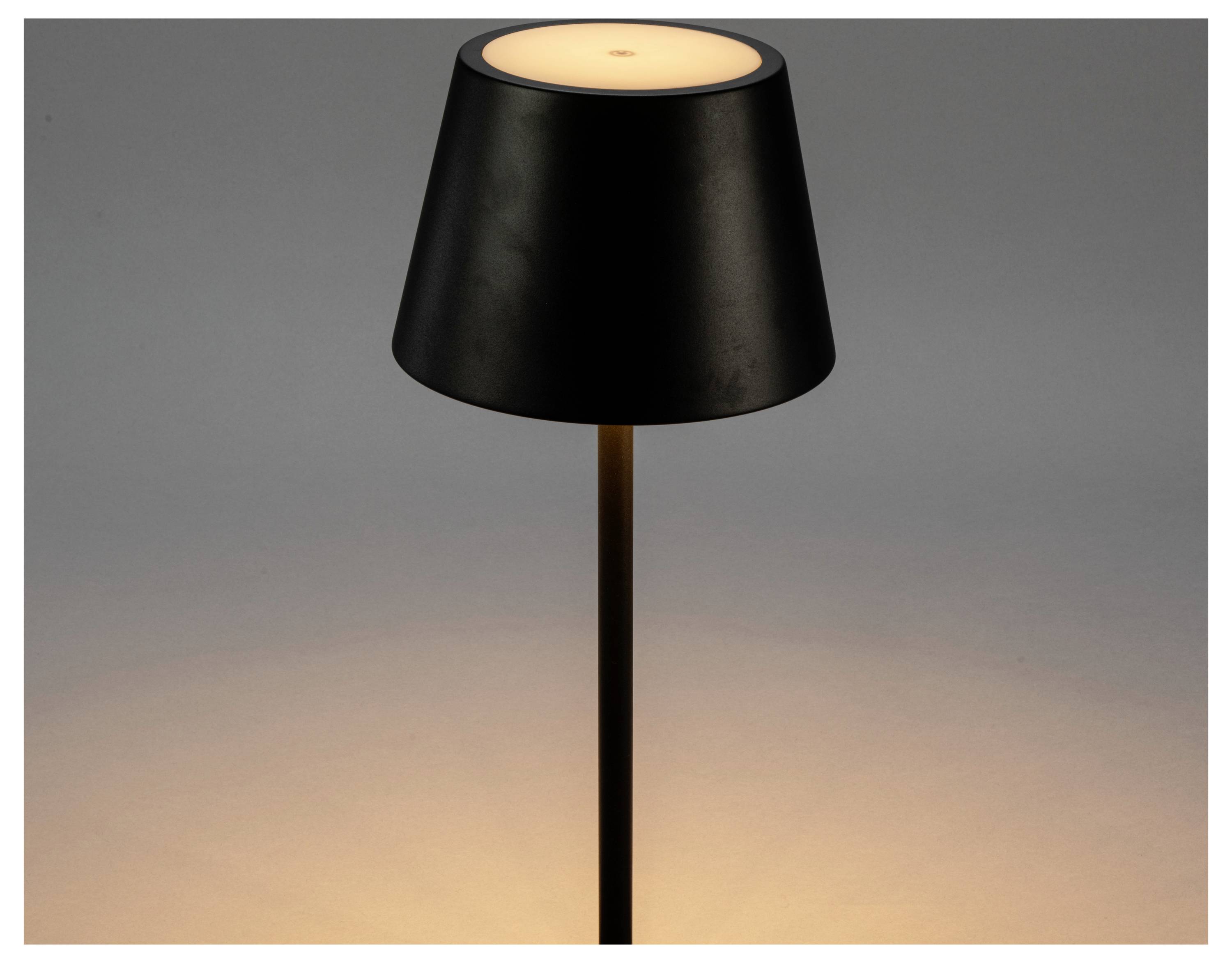 En svart, konisk bordslampa sänder ut ett varmt ljus och skapar ett mjukt sken på den omgivande ytan.