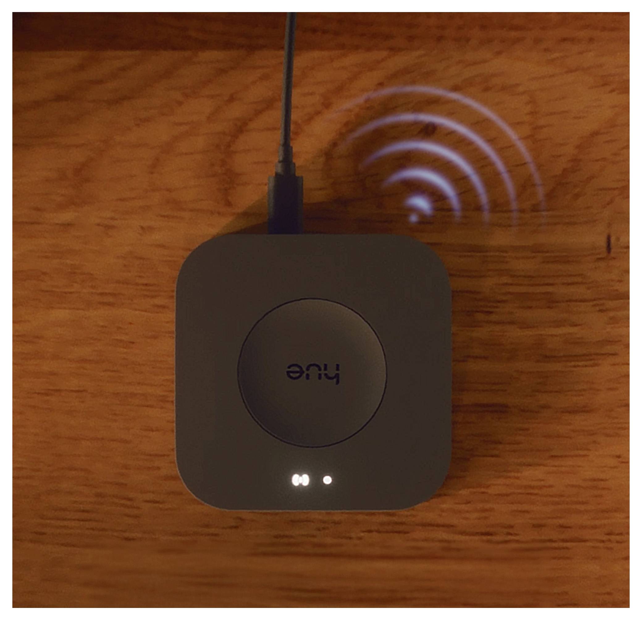 En liten svart fyrkantig smart-enhet märkt 'hue' ansluten via kabel, med två indikatorlampor på en träyta.