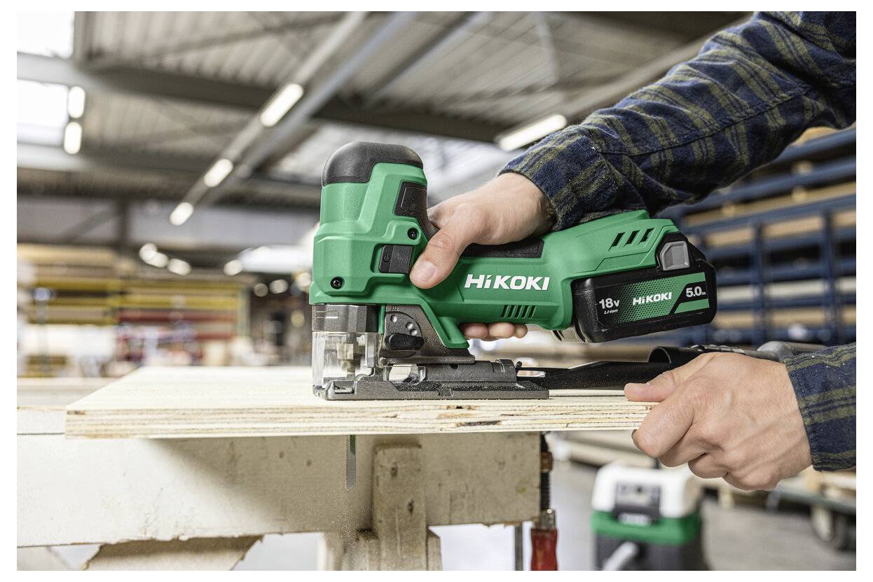 En person använder en grön Hikoki sticksåg för att skära igenom en träpanel i en välbelyst verkstad och visar därigenom dess precision och kraft.