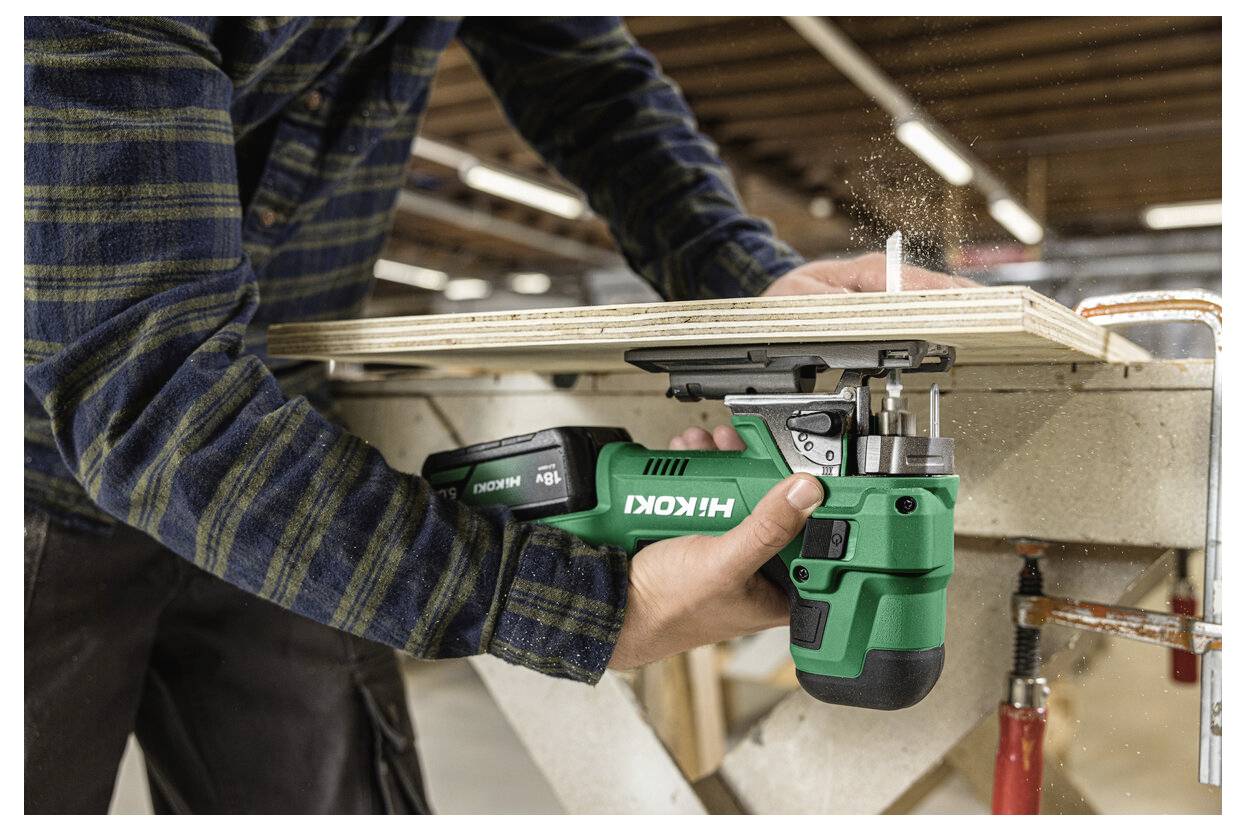 En person använder en grön Hikoki sladdlös såg för att såga igenom plywood på en arbetsbänk i en verkstad. Dammpartiklar syns i luften.