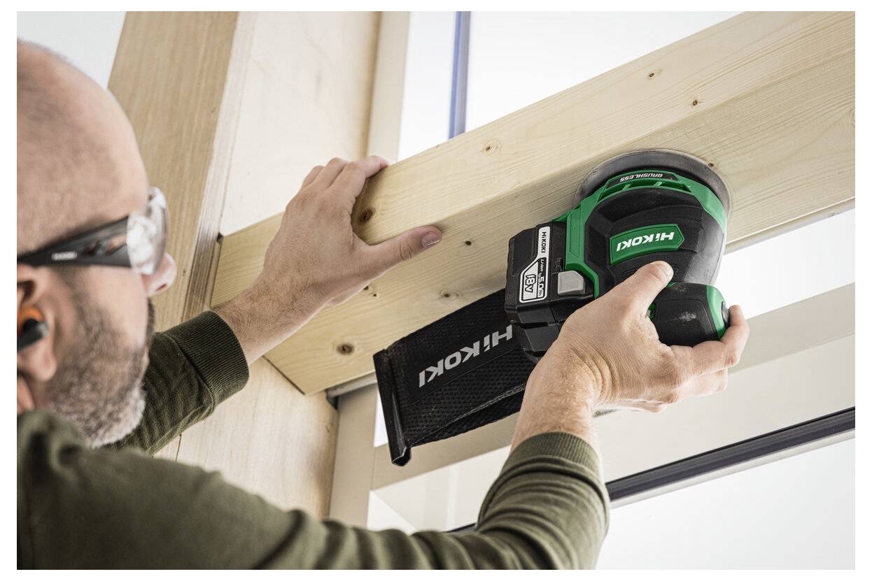 En person som bär skyddsglasögon och hörselskydd använder en grön Hitachi-slipmaskin på en träbalk inomhus.