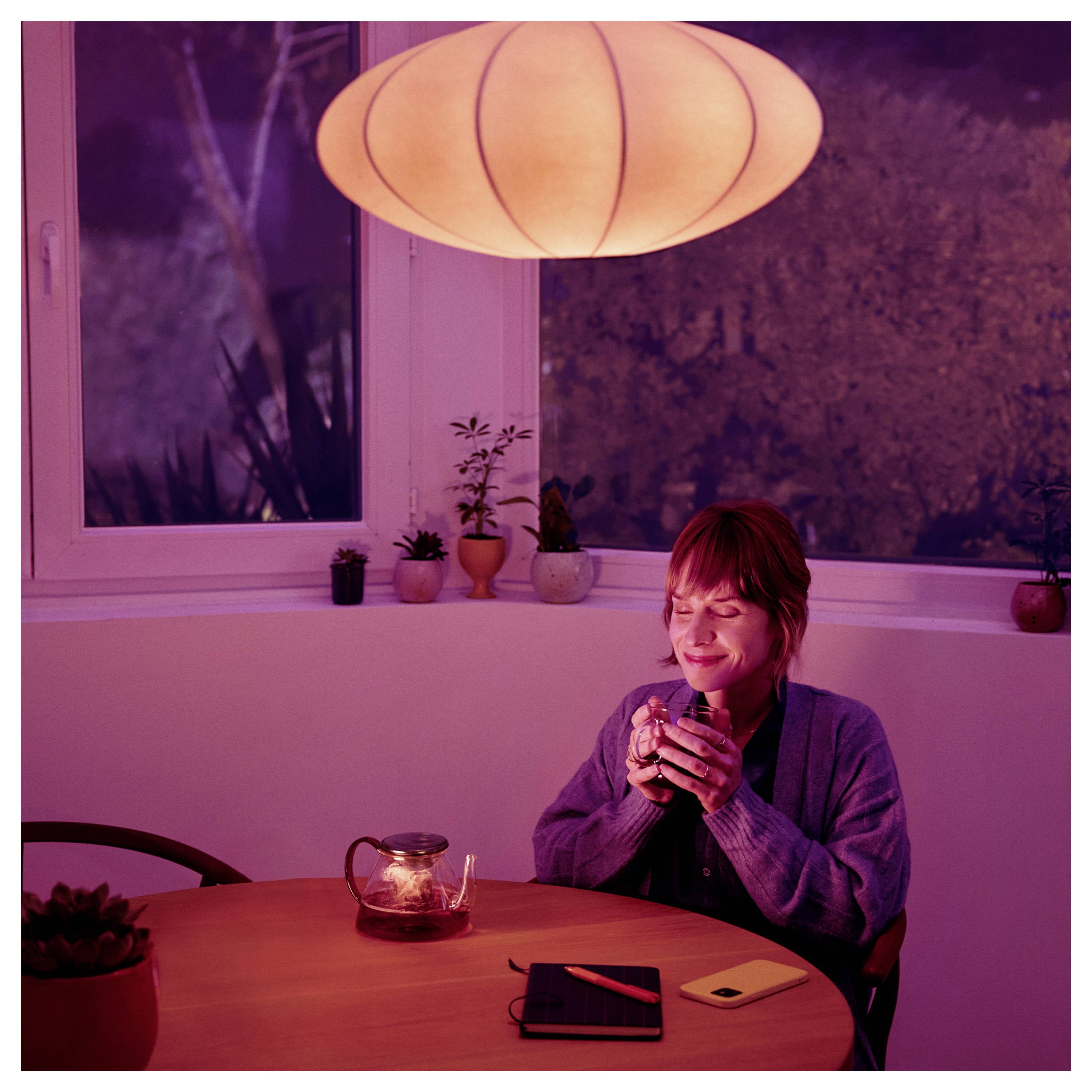 En person sitter vid ett bord i ett varmt upplyst rum, håller i en mugg och ler. En tekanna, en journal och ett kort ligger på bordet. En rund lampa hänger ovanför.