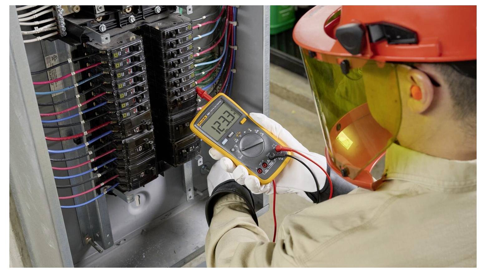 En tekniker i skyddsutrustning använder en multimeter för att kontrollera spänningen i en elcentral, där skärmen visar '123'.