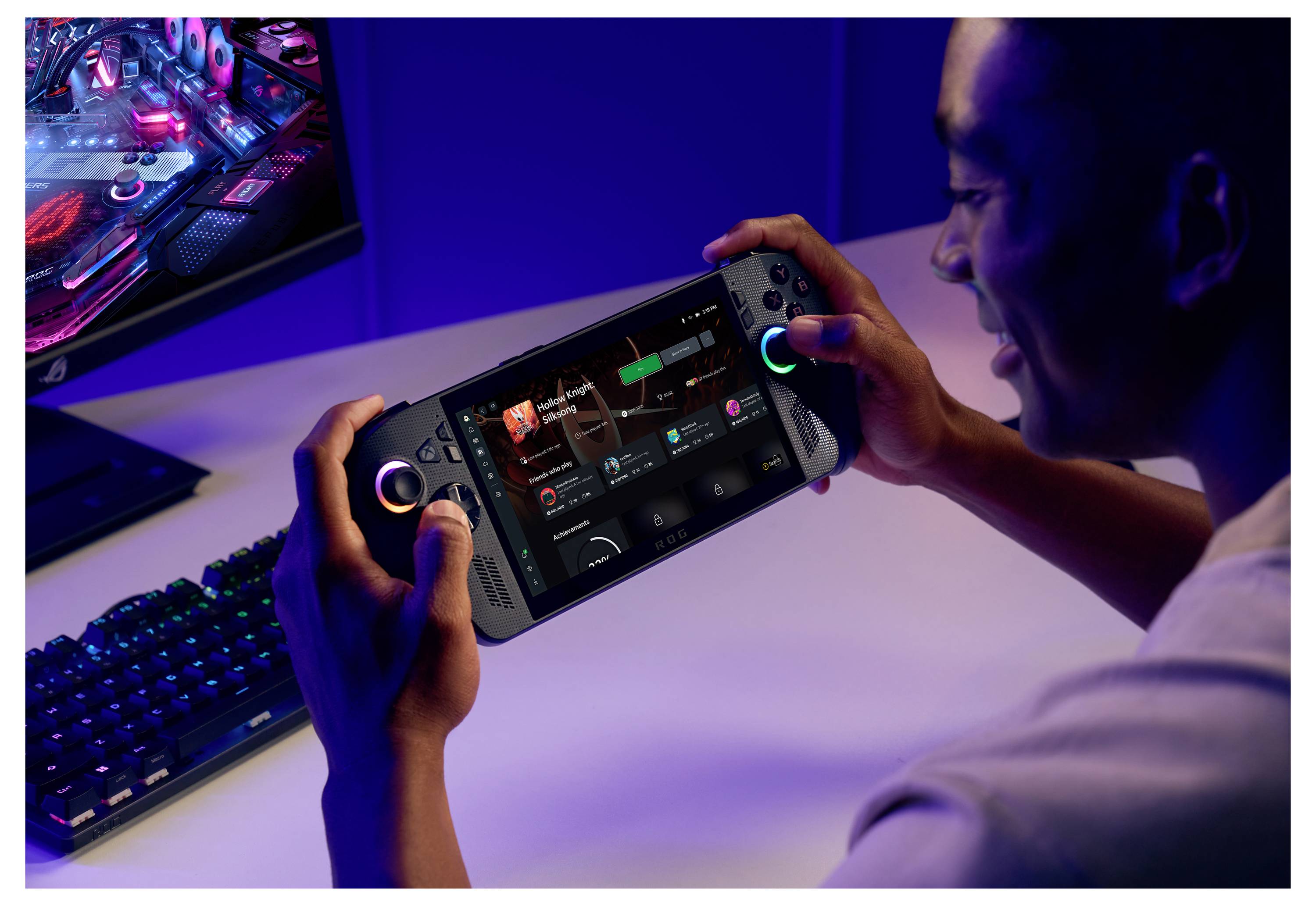 En person som spelar ett spel på en handhållen spelapparat, ler och sitter vid ett skrivbord med en datorskärm och tangentbord i närheten.