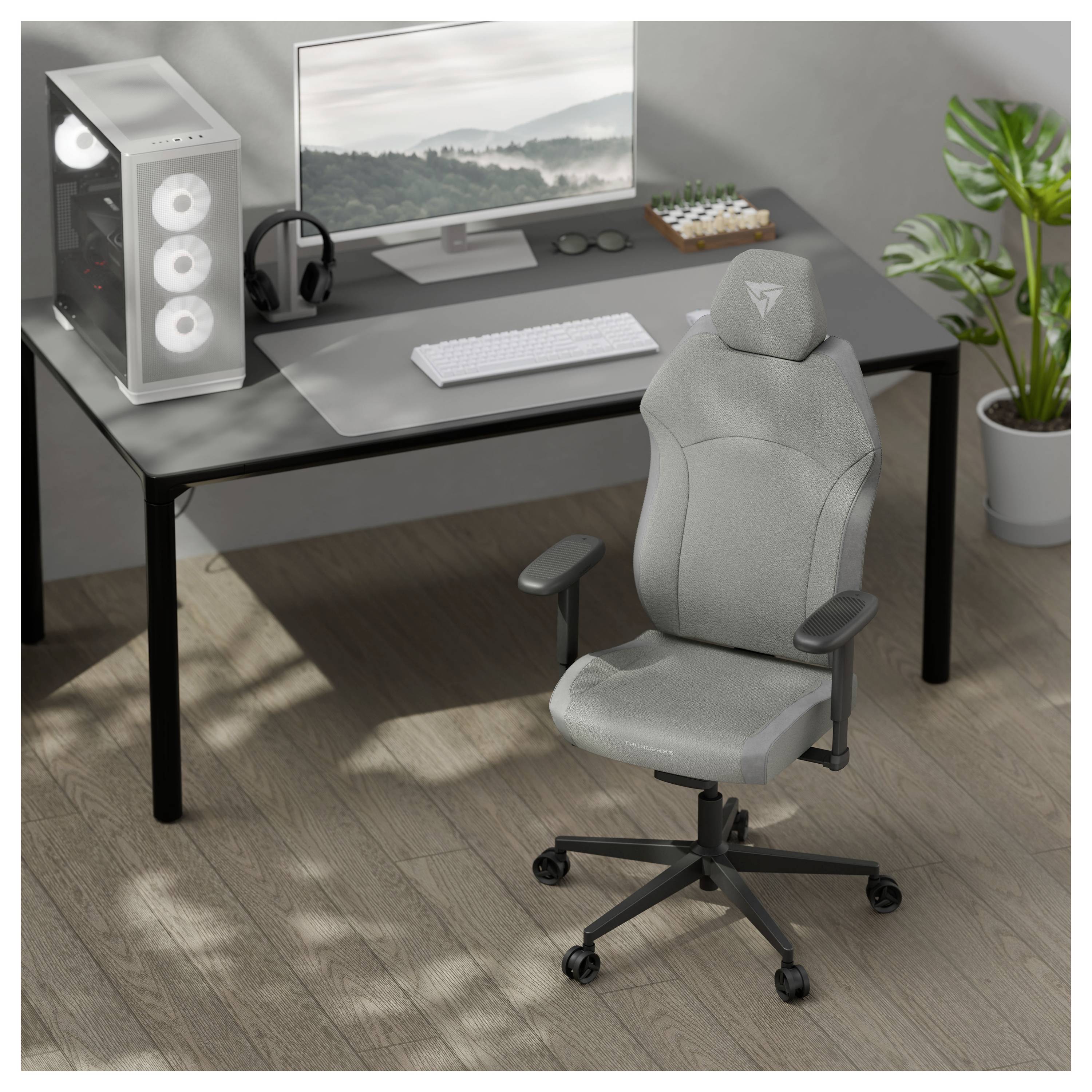 En modern kontorsmiljö med en grå gamingstol, ett skrivbord med en datorskärm, tangentbord, mus och ett vitt datorhölje bredvid en krukväxt.