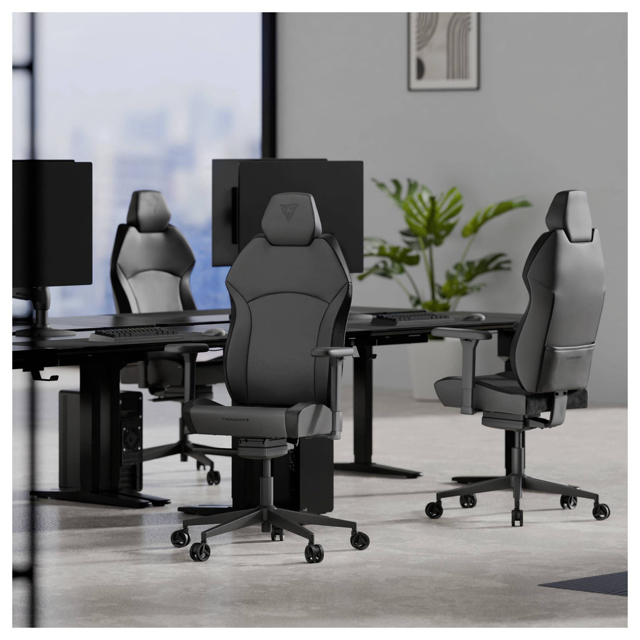 Ett modernt kontorsrum med tre svarta ergonomiska stolar, var och en placerad vid ett skrivbord med en datorskärm. En växt pryder bakgrunden.