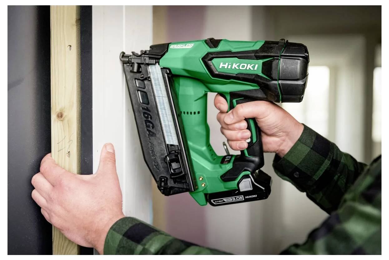 En person använder en grön och svart spikpistol för att fästa ett trästycke på en vägg. Verktyget är märkt 'HiKOKI' och har ett magasin.