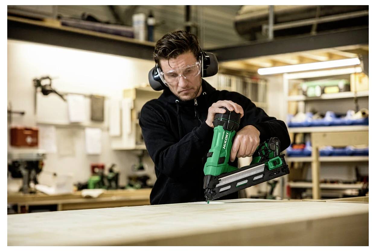 Person i en verkstad som använder en grön spikpistol på en träyta, bär skyddsglasögon och hörselskydd, fokuserad på uppgiften.