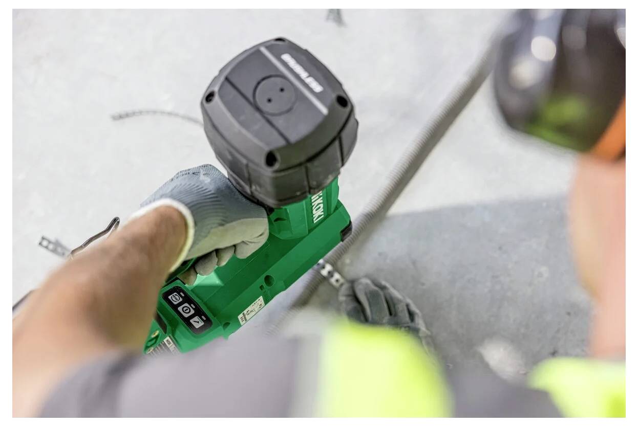En person i handskar och skyddsutrustning hanterar ett grönt elverktyg på en betongyta, vilket indikerar ett byggnads- eller reparationsarbete.