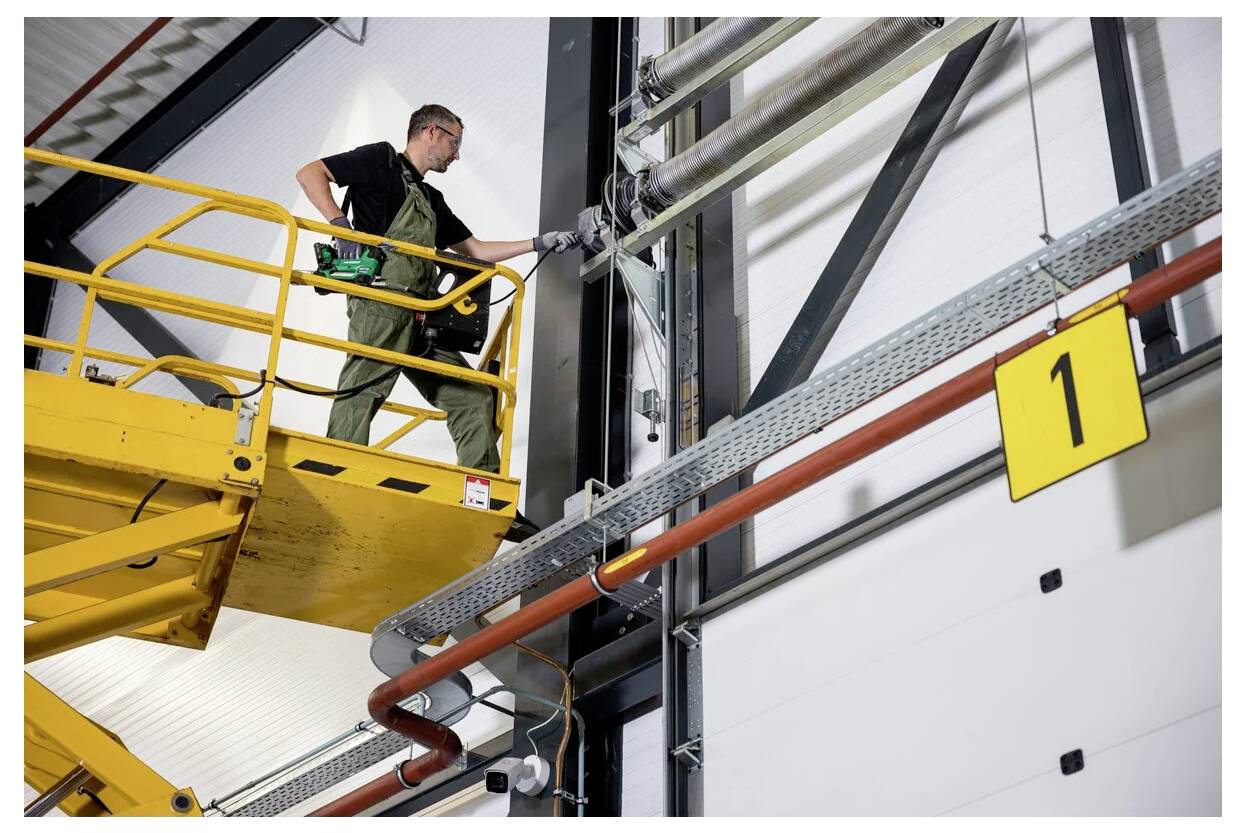 Arbetare i skyddsutrustning på en gul lyftkran som utför underhåll på rör i en industriell miljö, med en skylt märkt med nummer 1 i närheten.