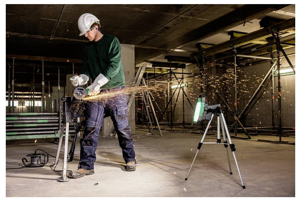 En byggnadsarbetare iklädd skyddsutrustning använder en vinkelslip på en metallbalk i en delvis uppförd konstruktion med byggnadsställningar runt omkring.