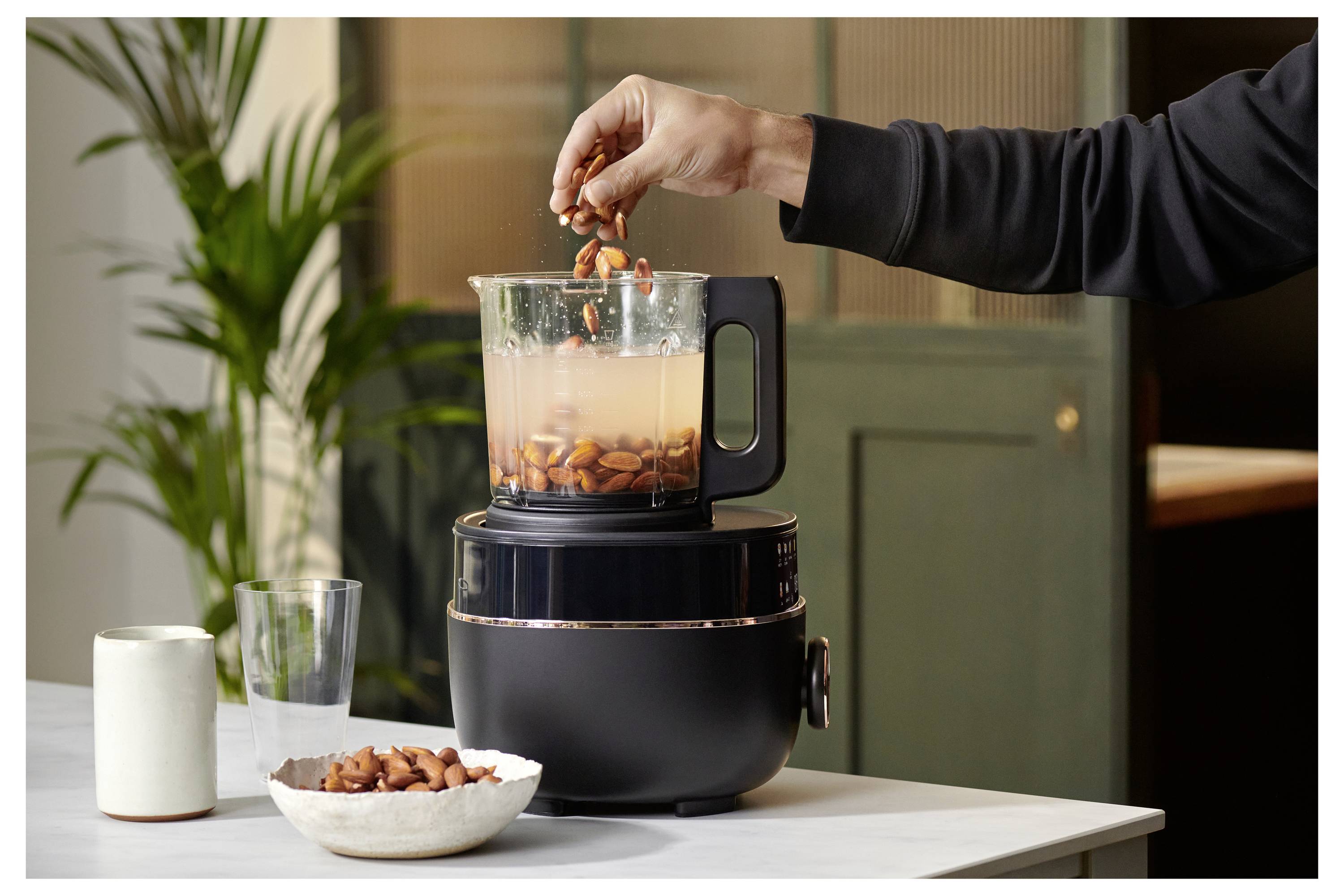 En person häller mandlar i en mixer fylld med vatten på ett köksbord. En kruka och köksredskap syns i bakgrunden.