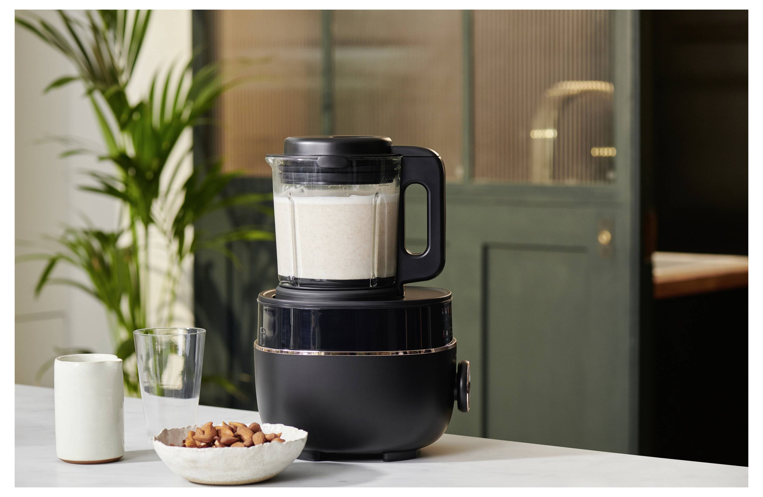 En svart mixer på en köksräknare fylld med en vit blandning, intill en skål med mandlar och ett glas, med en växt i bakgrunden.