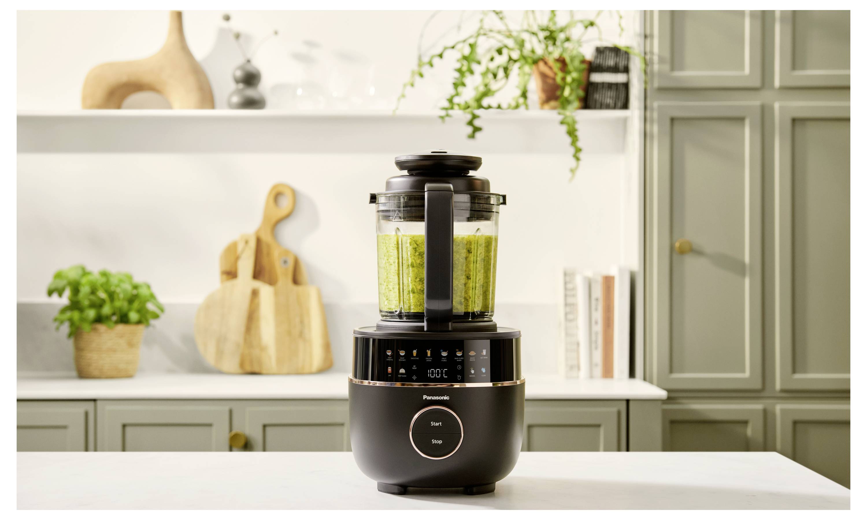 En blandare står på en köksbänk och blandar gröna ingredienser. I bakgrunden finns träredskap, växter och böcker.