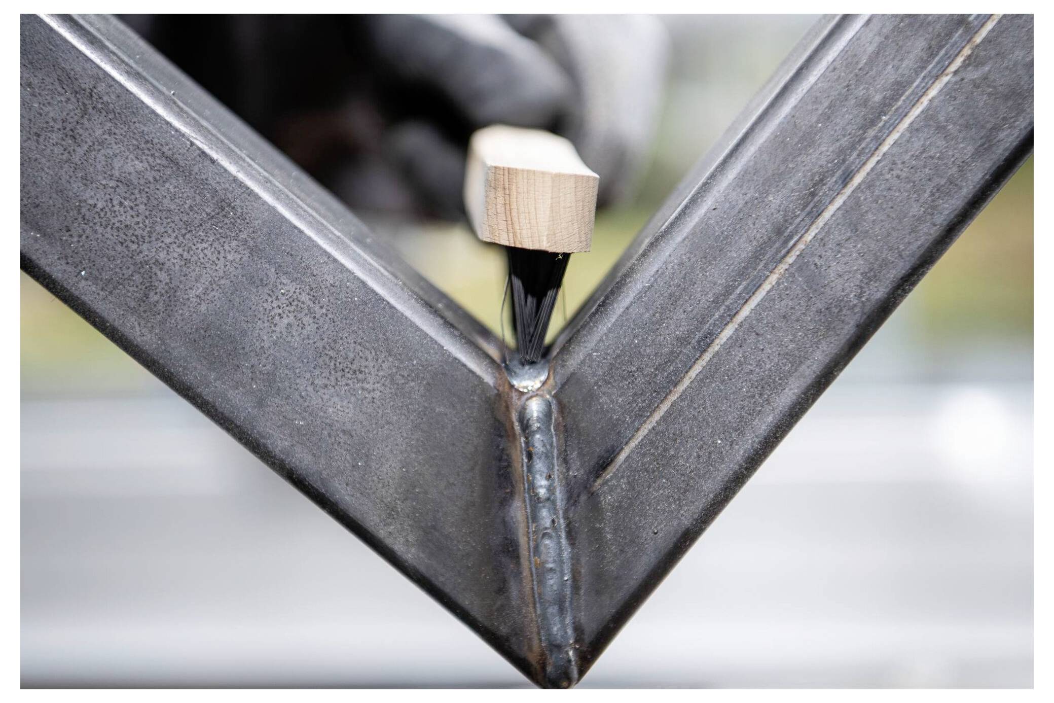 Närbild av en hand som använder en träborste för att applicera ett ytskikt på svetsfogen på en metalldetalj, vilket belyser precision i hantverksarbetet.