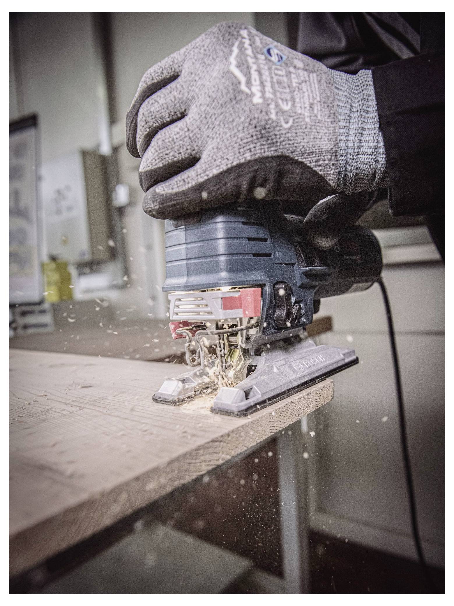 En person som bär handskar använder en sticksåg för att såga ett träplank, med sågspån som flyger runt, i en verkstadsmiljö.