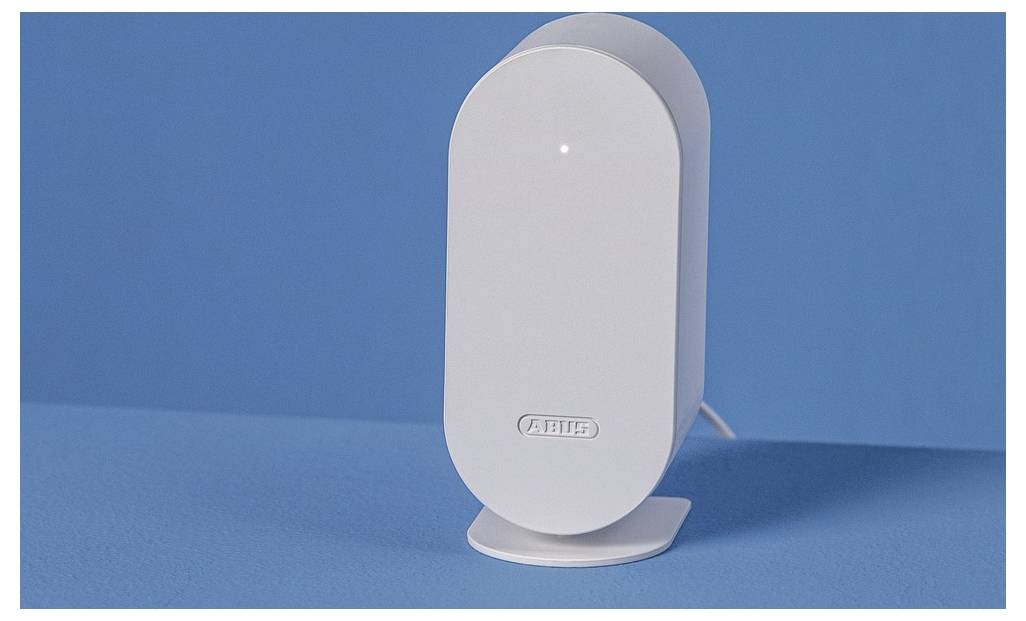 En vit, modern elektronisk enhet med ett litet ljus på toppen, placerad mot en blå bakgrund. Varumärket 'ABUS' syns på framsidan.